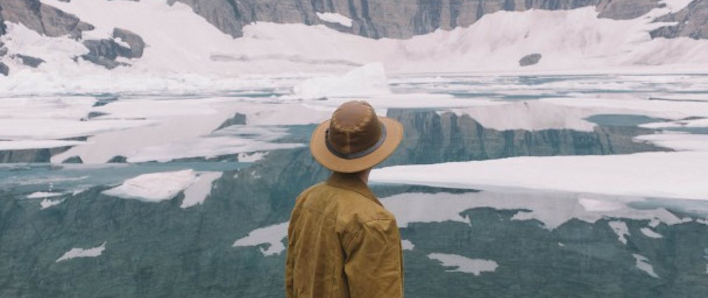 man in brown stetson and coat looks out at glacial pond with ice floats and sheer rock faces with snow