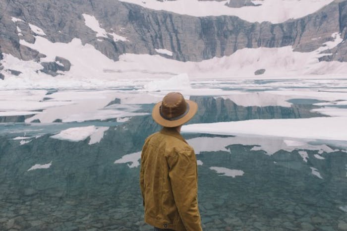 man in brown stetson and coat looks out at glacial pond with ice floats and sheer rock faces with snow