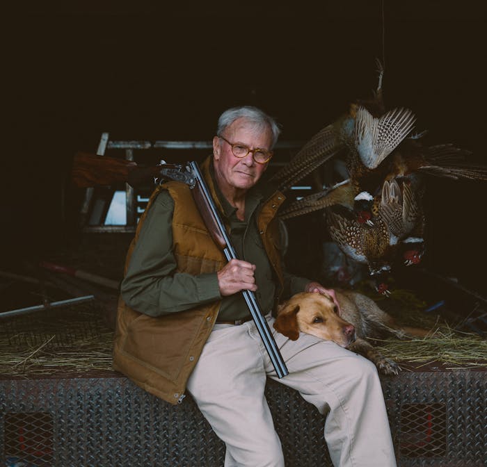 Tom Brokaw sits next to taxidermy pheasant and his hunting dog with a shotgun over his shoulder