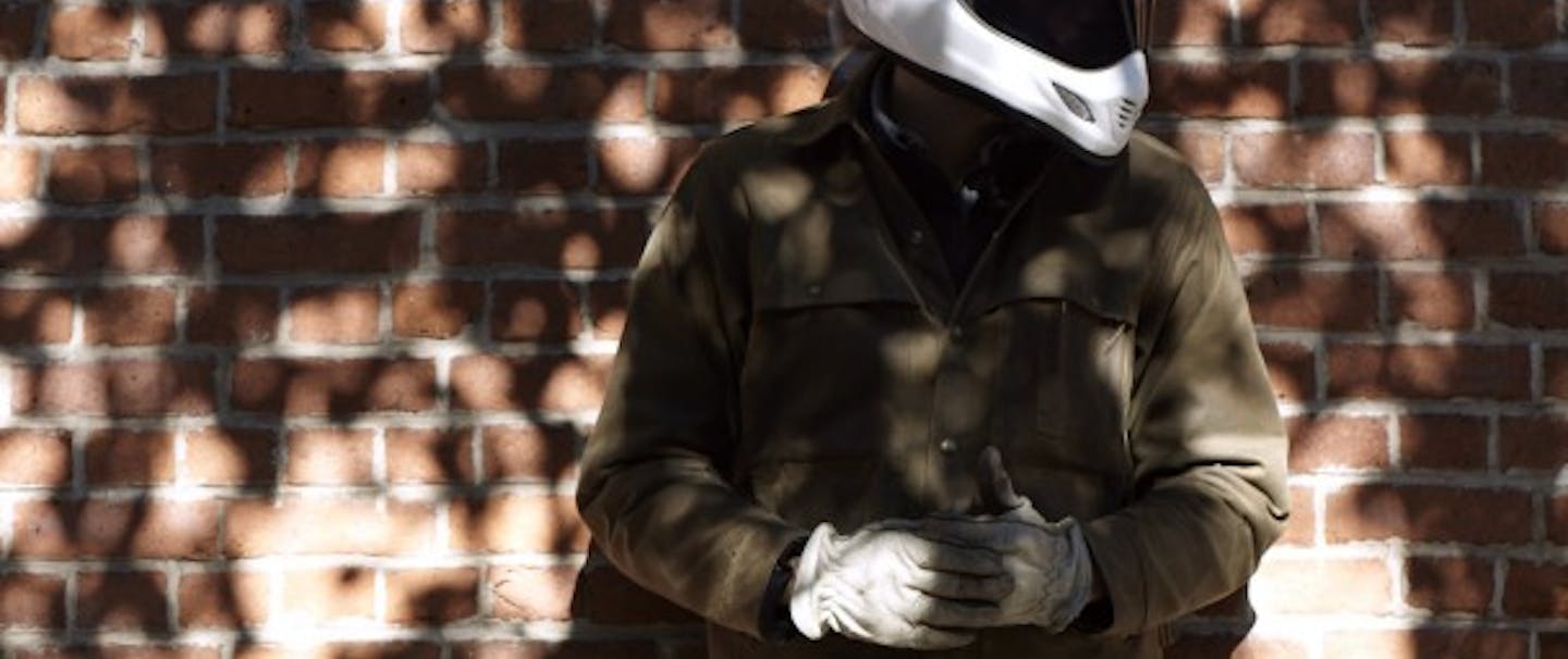 person in white motorcycle helmet with green filson jacket and white gloves in front of brick wall