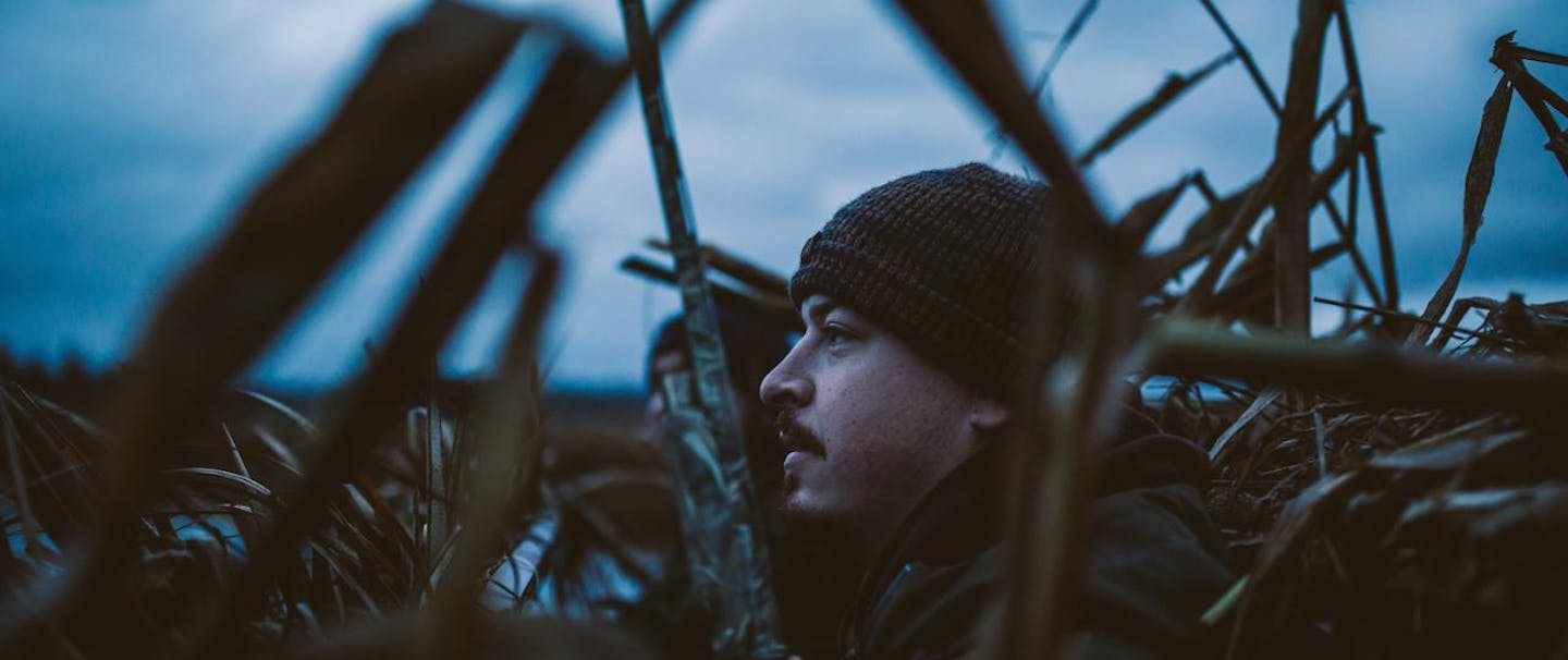 Filson Life - Winter Duck Hunting, hunters stand among reeds holding camouflage shotgun