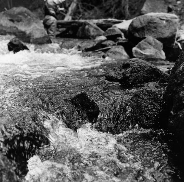 man stands upriver in rushing water among large rocks
