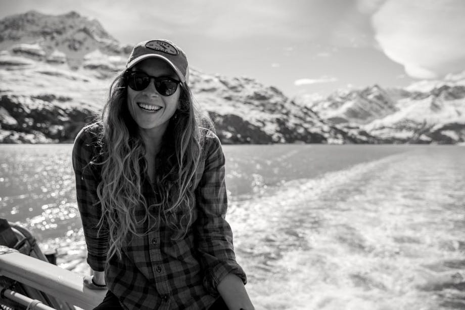woman on boat in sunglasses and hat and plaid shirt with snowy mountains in background