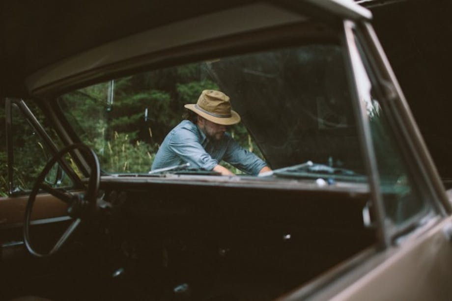 man in denim shirt and flat-brimmed hat works on hood of classic car