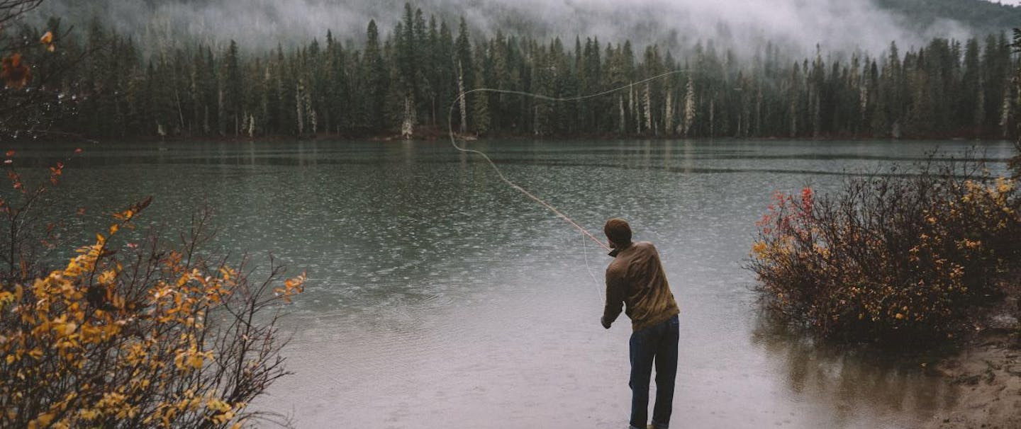 fly fisher casts into lake with pine trees and low lying clouds
