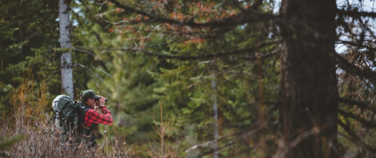 man in woods looks through binoculars wearing backpack and red flannel