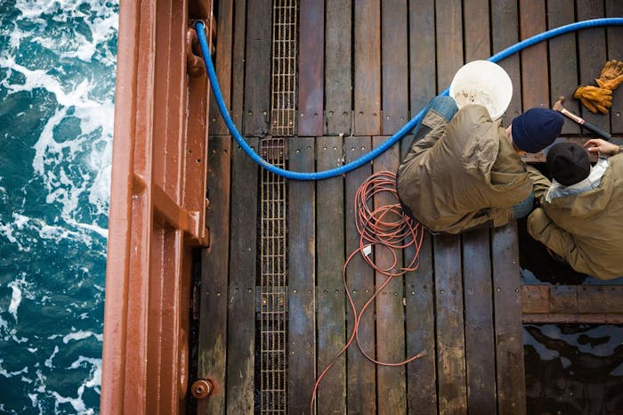 men in rain-proof brown jackets perform maintenance work on wooden deck at sea