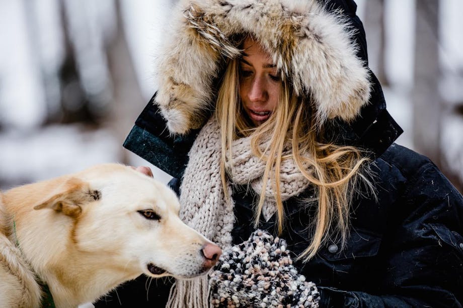Filson Life - Iditarider. woman with fur lined hooded parka and knit scarf pets white dog