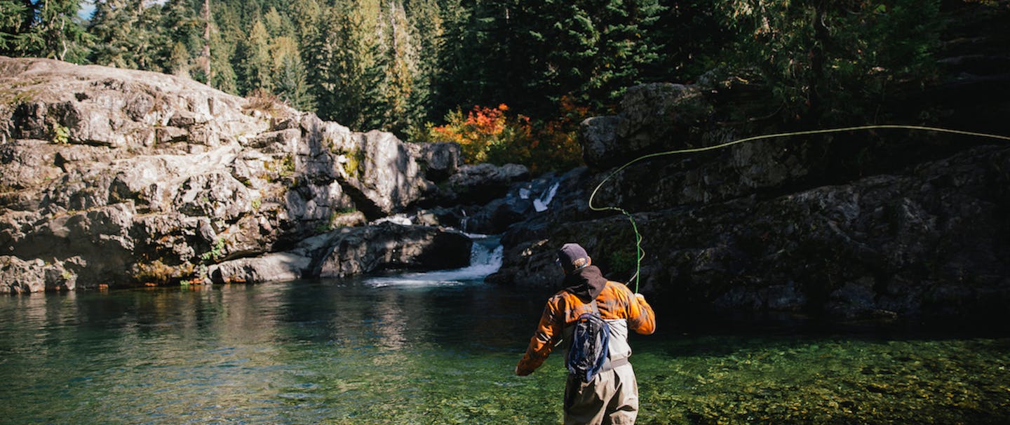 Man casting fly-fishing rod in clear water with waterfall and alpine ridge backdrop