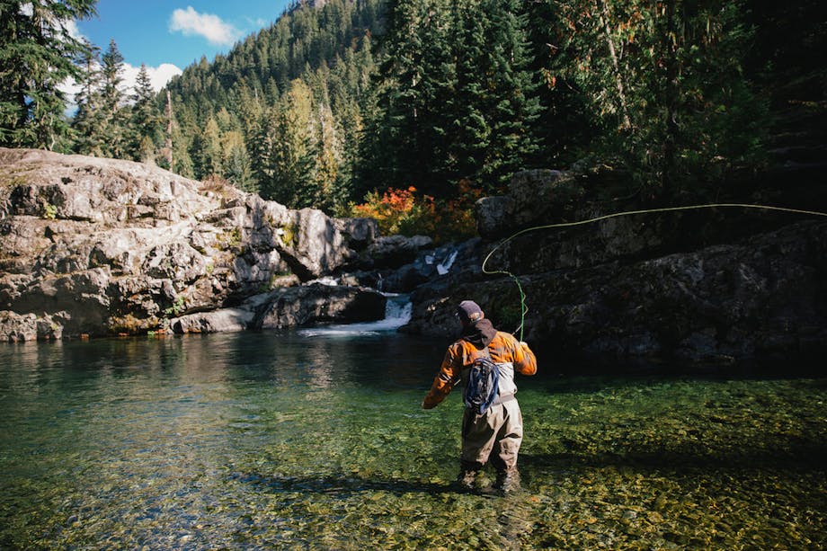 Man casting fly-fishing rod in clear water with waterfall and alpine ridge backdrop
