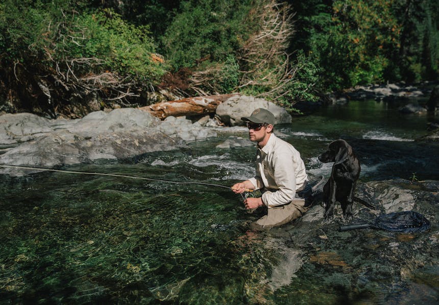 Man crouched in the middle of an alpine stream fly fishing alongside a dog to his right.