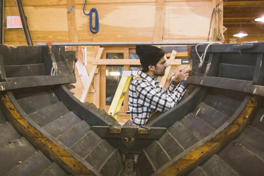 man in plaid shirt and black beanie works on hull of small wooden boat