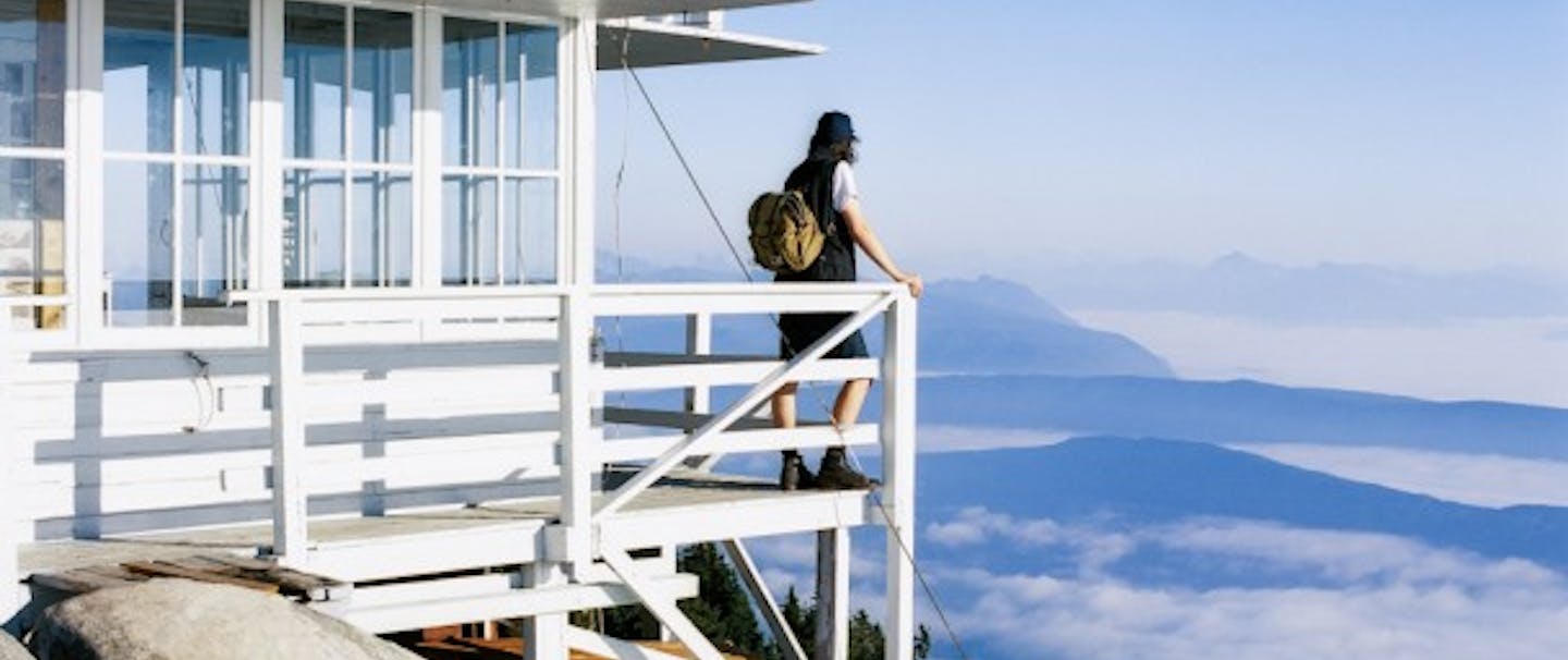 person with filson pack stands on deck of high mountain lookout above clouds