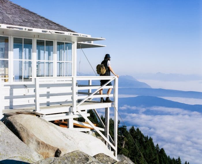 person with filson pack stands on deck of high mountain lookout above clouds