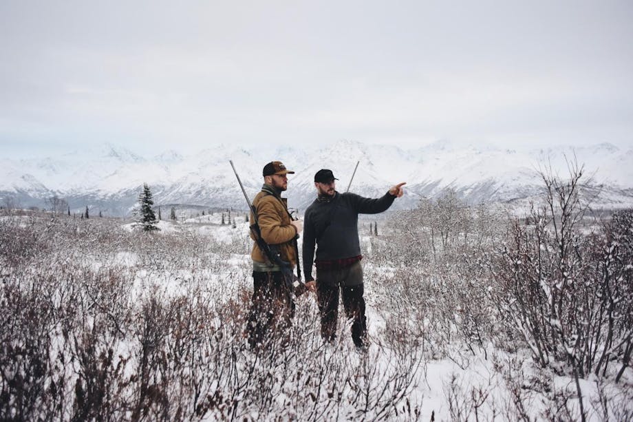 Two hunters with rifles point and look into the distance in a snowy field
