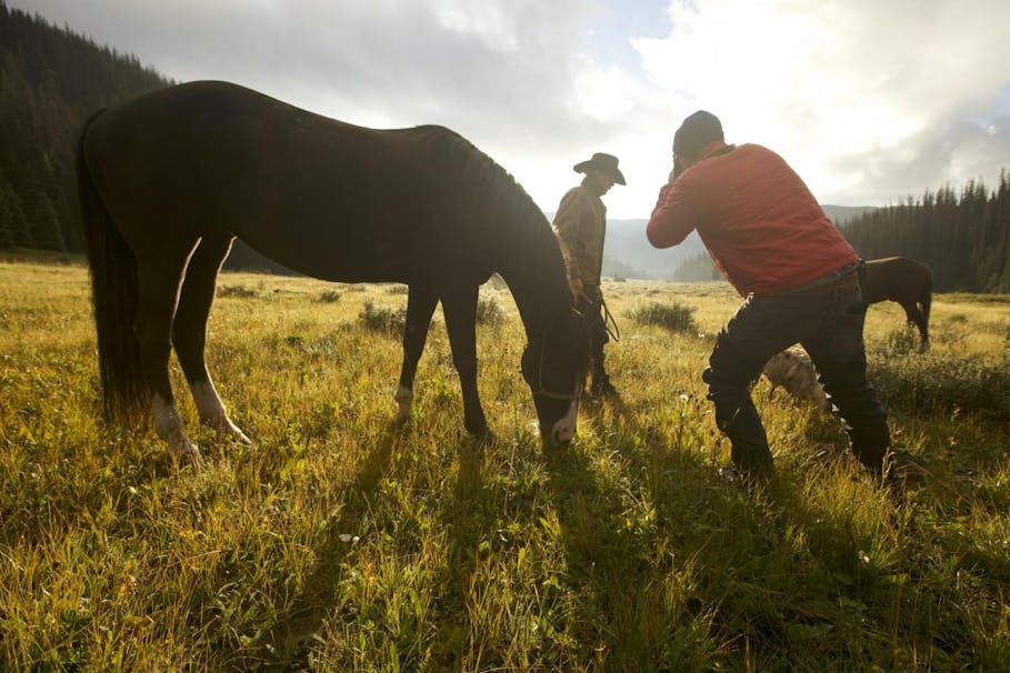 man takes picture of cowboy next to unsaddled horse forest clearing