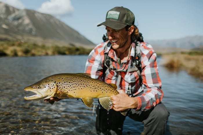 fisherman holding large fish kneeling in river wearing red and white flannel and green trucker hat