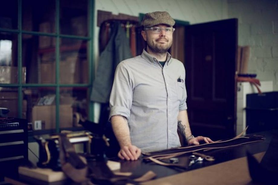 man in baker boy hat and buttondown shirt stands at leather goods workbench