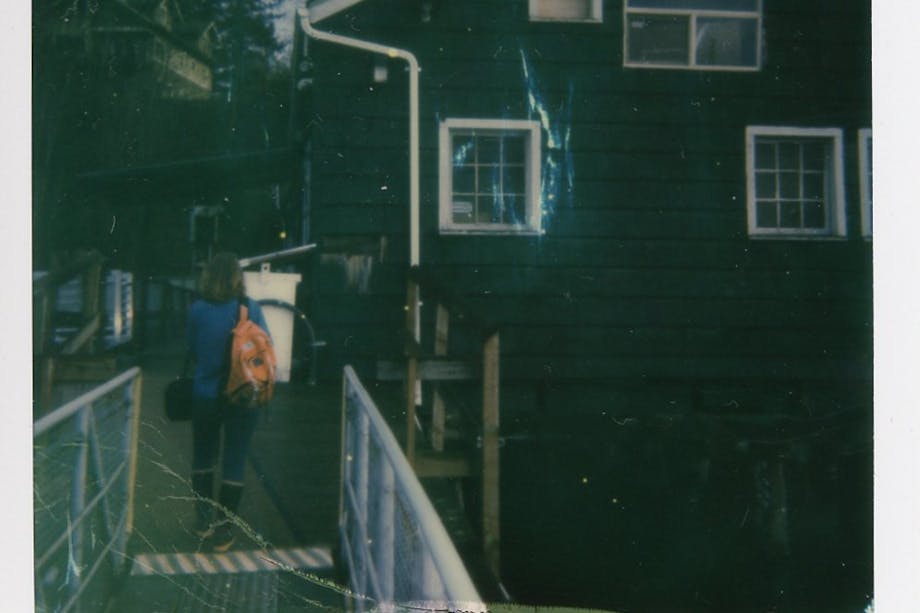 polaroid; woman stands on walkway leading to green house with three windows