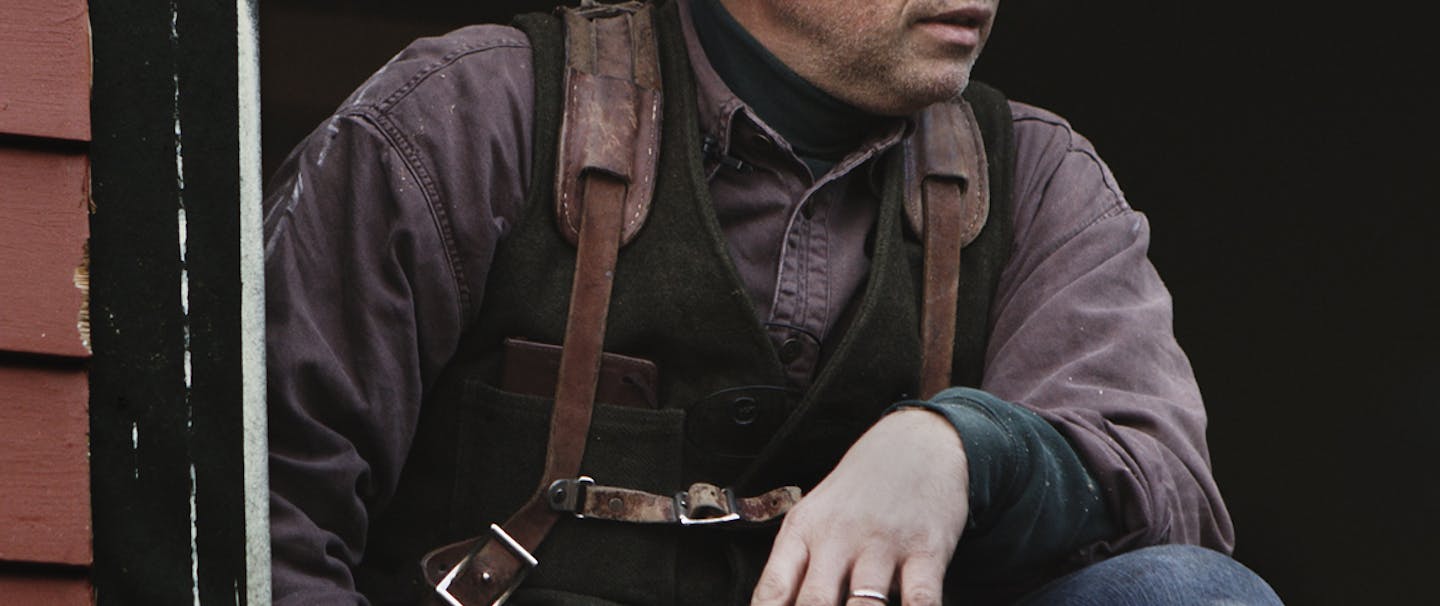 Daniel Westbrook kneels with grey newsboy cap, black vest, purple buttonup and leather holster straps