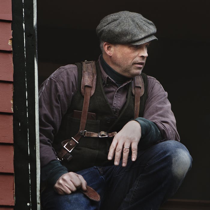 Daniel Westbrook kneels with grey newsboy cap, black vest, purple buttonup and leather holster straps