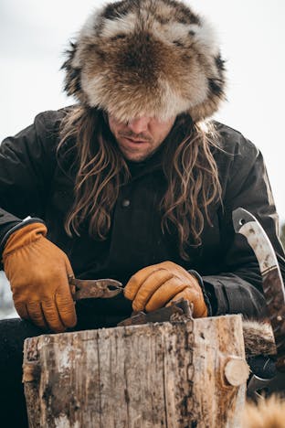man with long brown hair, wearing a fur hat, black wool jacket and goat skin gloves working with tools on top of a whole log of wood