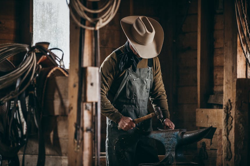 a man wearing black work overalls, a dark brown long sleeve and white cowboy hat looking down as he works on a horshoe in a barn