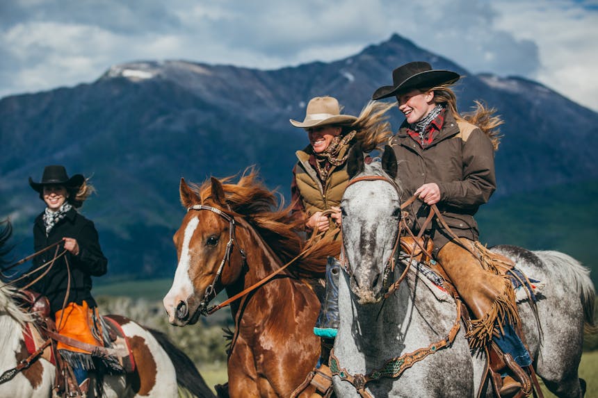 three women wearing jeans, chaps and cowboy hats riding their horses with snowcapped mountains in the background