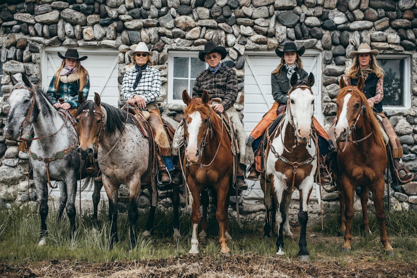 Five horses (two brown with white faces, one brown and white, one light brown with a darker face and one grey and black) with five riders wearing cowboy hats and flannels sitting in the saddle in a line in front of a stone building