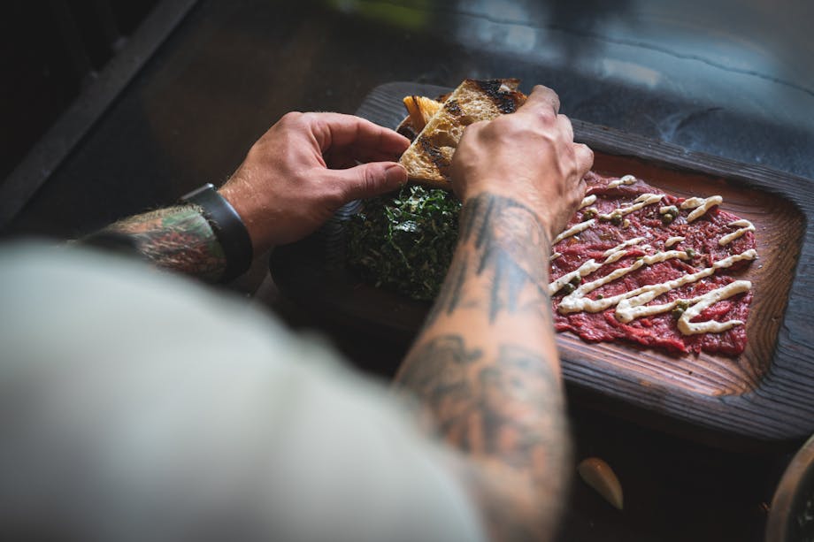 chef plating the dish with the final touches of drizzled with horseradish sauce, a side of kale salad and grilled bread on a dark wood serving dish