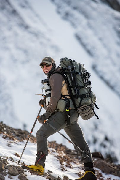 man wearing green hiking pack, dark green pants, shirt and cap, carrying a big camera and hiking poles up a snowy mountainside