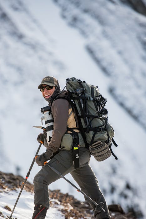 man wearing green hiking pack, dark green pants, shirt and cap, carrying a big camera and hiking poles up a snowy mountainside
