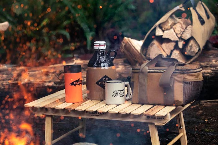 camping gear, orange mug, brown glass growler, cream ceramic mug, brown utility bag and a tan log carrier full of wood, in the woods next to a campfire