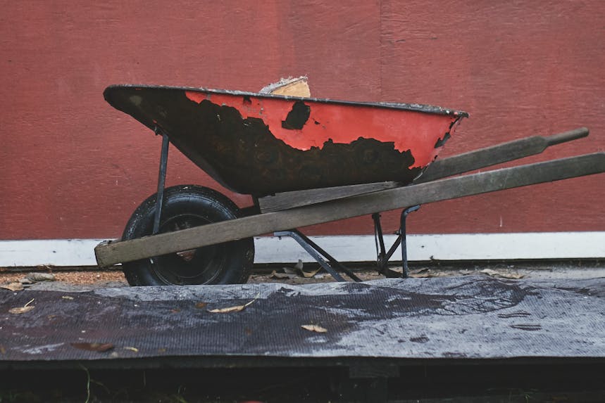 the side angle of a red wheel barrow with wood in it next to the base where it will be stacked in front of a red building