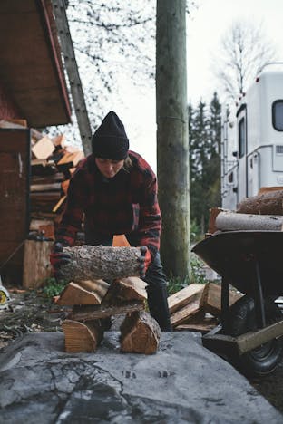 brunette woman wearing a black knit hat, red and black plaid sweater stacking firewood on a dark tarp