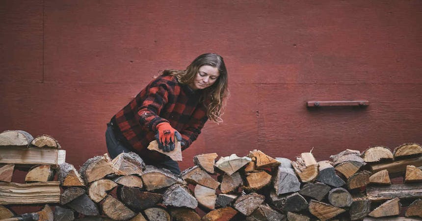 brunette woman wearing a red and black plaid sweater, black and orange gloves and dark pants stacking wood on a wood pile with a red building behind her
