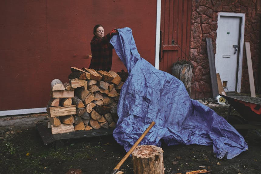 brunette women wearing a red and black plaid sweater pulling a faded blue tarp over the top of a stacked wood pile