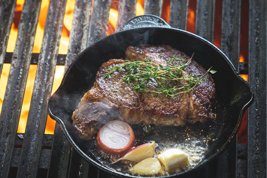 cast iron pan on a flaming outdoor grill with browned steak, halved red onion, whole garlic and sprigs of thyme