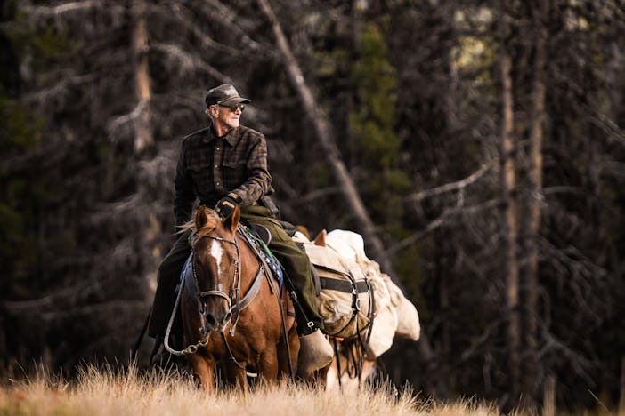 man wearing a brown flannel and dark ball cap riding a brown horse with cream saddle bags on a trail exiting a wooded area