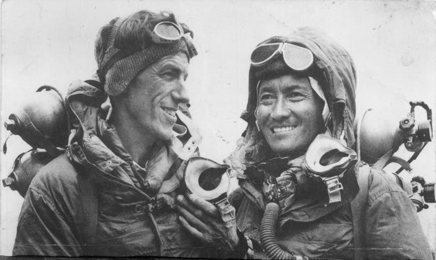 a black and white image of two mountaineering men with their googles on their foreheads, bundled up with jackets and wearing packs smiling from ear to ear