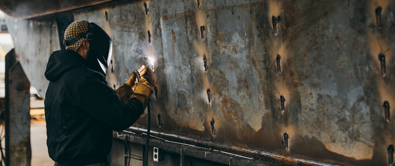man wearing brown beanie, black work jacket, jeans and protective gear as he welds a metal boat