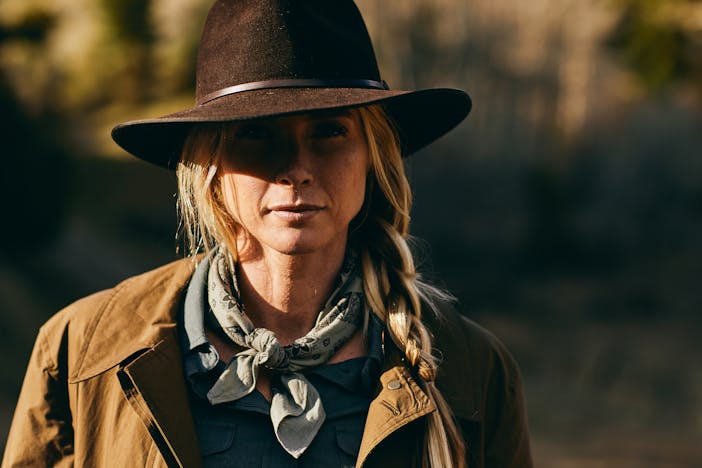 blonde woman with a long braid, grey scarf, brown cowboy hat and brown jacket with a blurry wooded background