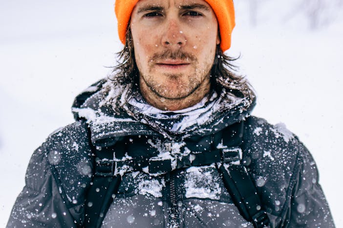 a man with a blaze orange beanie and dark grey down jacket with snow falling and collecting on his jacket and around his neck gaiter