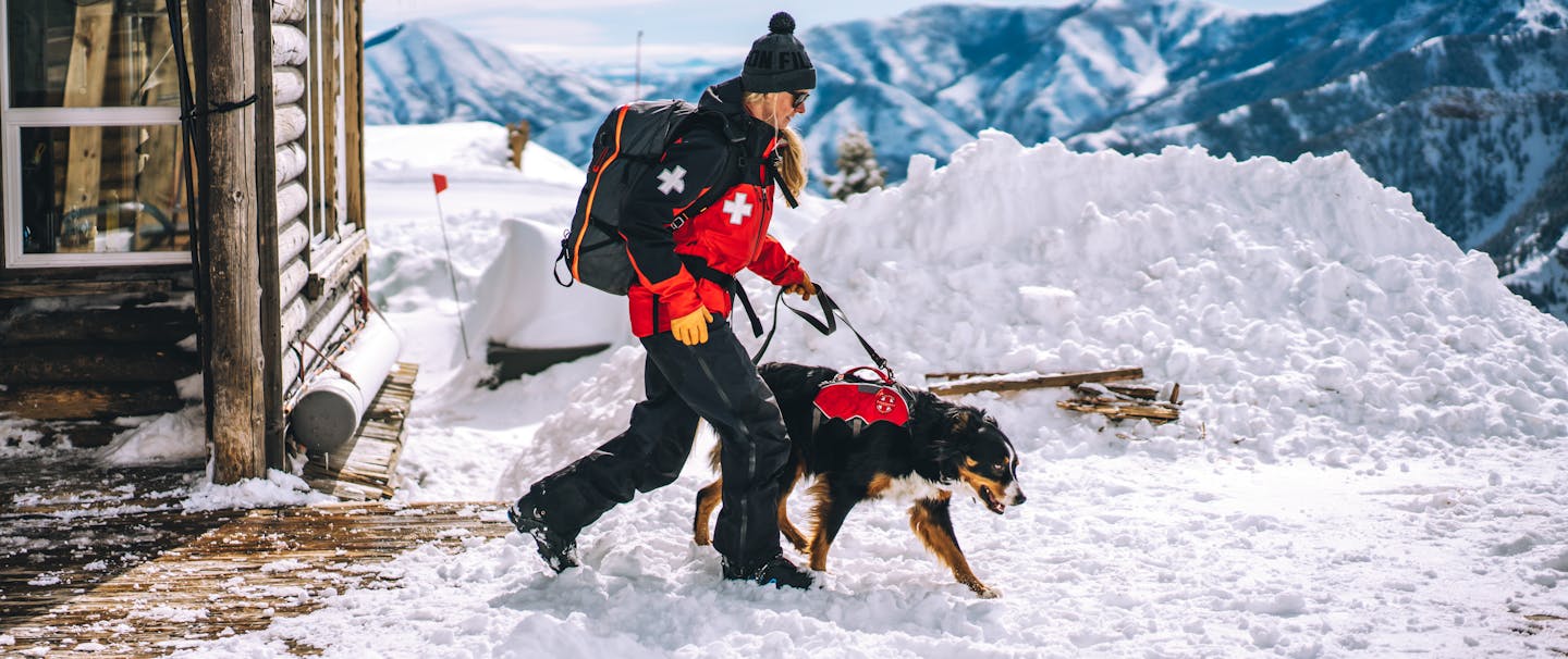 a blonde ski patroller wearing her red and black jacket with a white medical cross on the shoulder, black snow pants, black boots and holding the leash of her black, brown and white dog wearing his red and black rescue vest as they walk away from the chalet