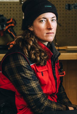 a brunette woman wearing a black beanie, red and black vest and brown and black plaid flannel sitting on a stool in a workshop glaring at the camera