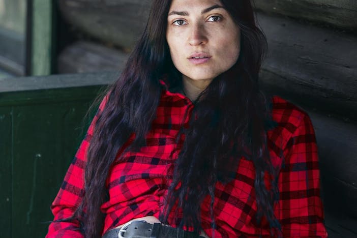 Native American woman leaning back in her chair on the porch of a wooden cabin wearing a black and red flannel shirt tucked into her black jeans and a black leather belt
