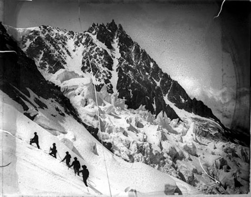 a black and white image of a mountain peak close in distance while five people climb the steep incline from afar making them look like ants