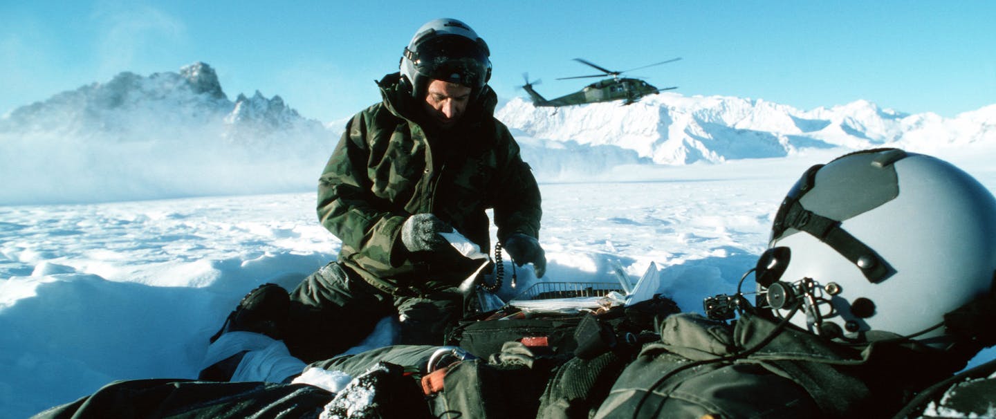 two Air Force men wearing green snowsuits, one sitting up, while the other lays down resting on a bag as a helicopter takes off in the snowy mountains in Alaska