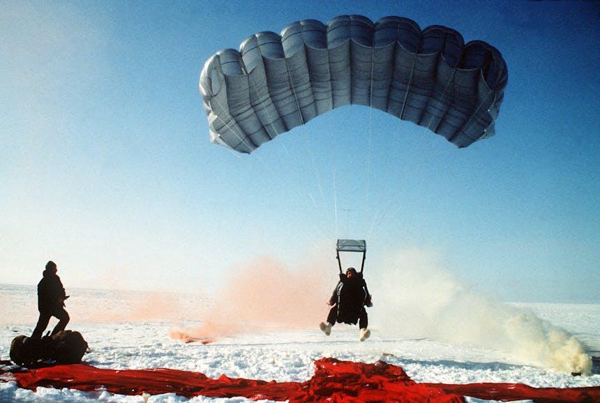 a historic photo of Air Force jump training, one man is about to land with his feet stretched out and his blue parachute open about 2 feet off the ground where there is a red landing strip and a yellow flare to the right and orange to the left while someone stands there to assist if needed