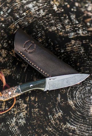 top down view of an outdoor knife with dark and colored wood handle and leather strap hanging from handle next to the leather sleeve on a tree stump